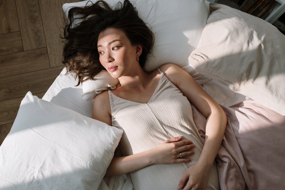 Asian woman in sleepwear lying on bed with hand on belly, enjoying morning sunlight.