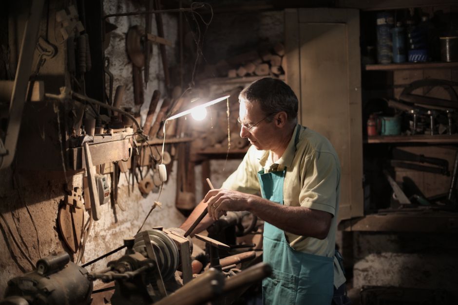 Focused artisan in apron and eyeglasses using lathe machine while holding metal detail in hand during working in dark workshop with lighting lamp under head