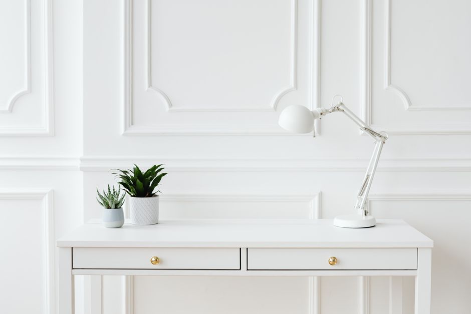 A minimalist white desk setup featuring potted plants and a sleek desk lamp against a white wall.